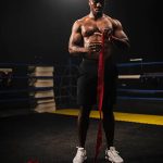 a muscular boxer stands in a dim gym, bending to tie a red ribbon